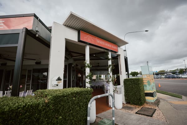 Entrance to White Horse Ranch showing outdoor covered area with tables and chairs set up, surrounded by greenery and a street view with parked cars and a painted utility box.