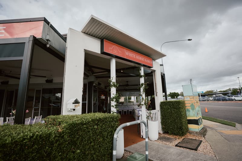Entrance to White Horse Ranch showing outdoor covered area with tables and chairs set up, surrounded by greenery and a street view with parked cars and a painted utility box.