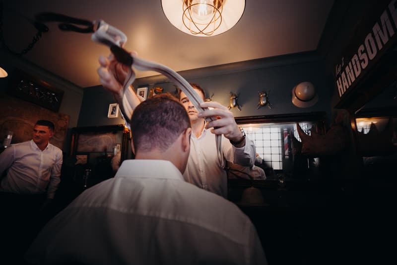 Groom Brent is assisted by a man putting on suspenders in a room with vintage decor, while another man in a white shirt stands nearby at Preston Peak Winery Functions Centre.