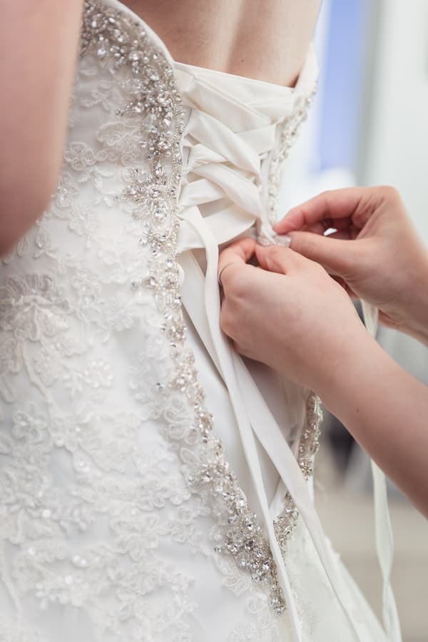 The bride's wedding dress is being laced up by another person, showing detailed beadwork and lace on the gown.