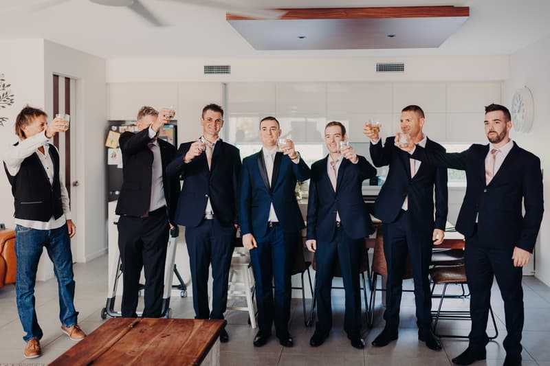 The groom Dylan and six groomsmen raise glasses in a toast indoors in a modern kitchen or dining area.