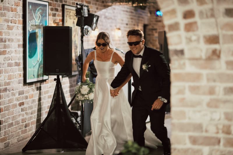 Bride Ashleigh and groom James wearing sunglasses hold hands and walk together inside The Cellar at Sandstone Point Hotel during the reception.
