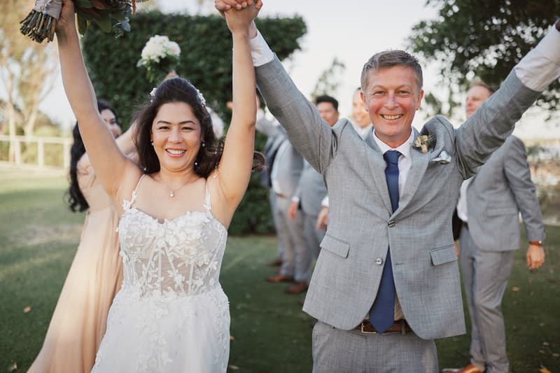 Bride Jheng and groom Dane celebrate outdoors at Sandstone Point Hotel with bridal party members in the background.