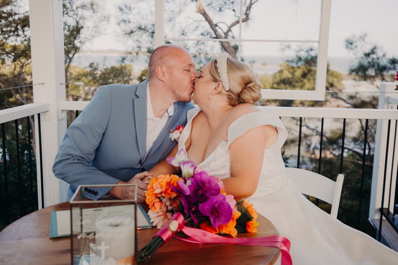Jacquelyne and Arran share a kiss at a table during their wedding ceremony at Sandstone Point Hotel — Pavilion, with a bouquet and a candle on the table.