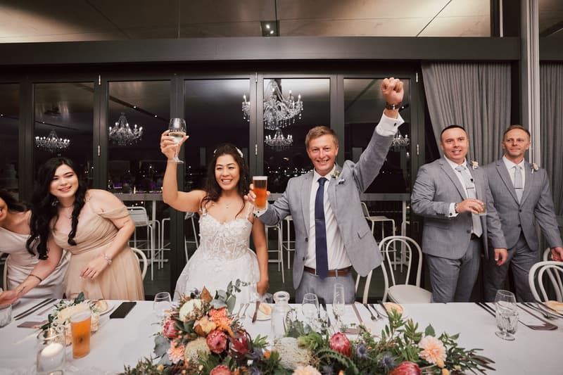 The bride Jheng and groom Dane raise their glasses in a toast at the reception in The Pumicestone Room, Sandstone Point Hotel, joined by bridesmaids and groomsmen standing behind the head table decorated with floral arrangements.
