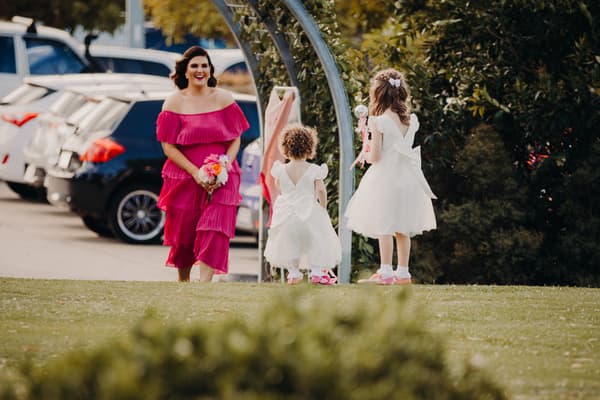 A bridesmaid in a bright pink dress holding a bouquet walks toward two flower girls in white dresses standing near a metal arch at Sandstone Point Hotel — Pavilion.