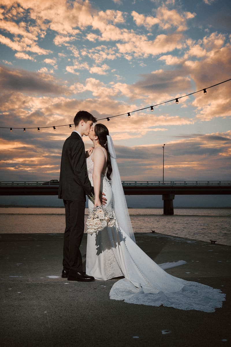 Terion and Tyler share a kiss during a styled portrait session, with string lights overhead and a bridge in the background. Getting ready photos by She Said Yes.