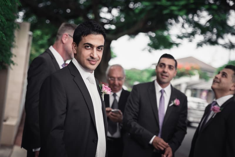 The groom Pasha stands with groomsmen outside under trees, all wearing dark suits with pink boutonnieres.