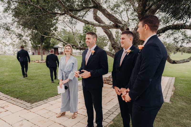 Three groomsmen wearing dark suits with peach boutonnieres stand on a paved area near a tree at Preston Peak Winery Functions Centre, accompanied by a woman in a light blue dress holding a clutch. Two other men in dark suits walk away on the grass in the background.