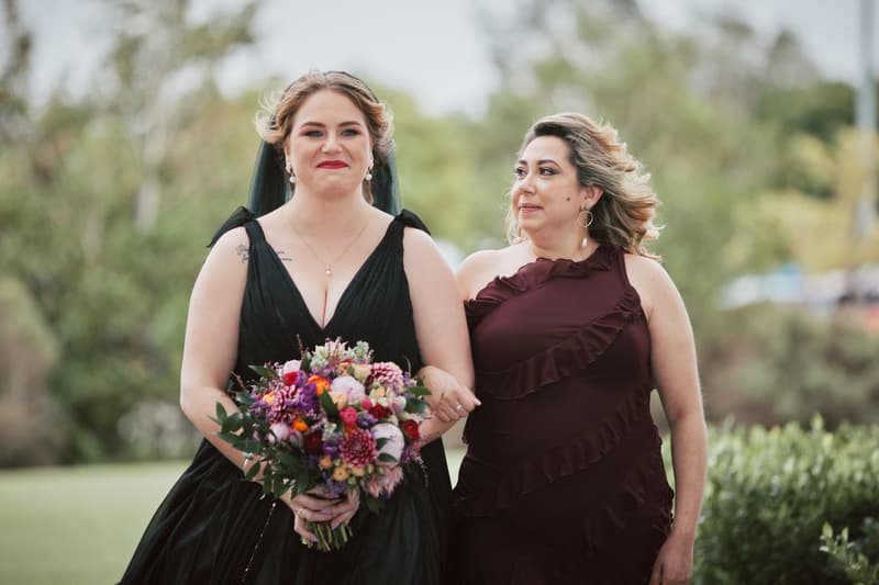 The bride in a black dress holding a colorful bouquet walks arm-in-arm with a woman in a burgundy dress outdoors at Sandstone Point Hotel — The Pavilion.
