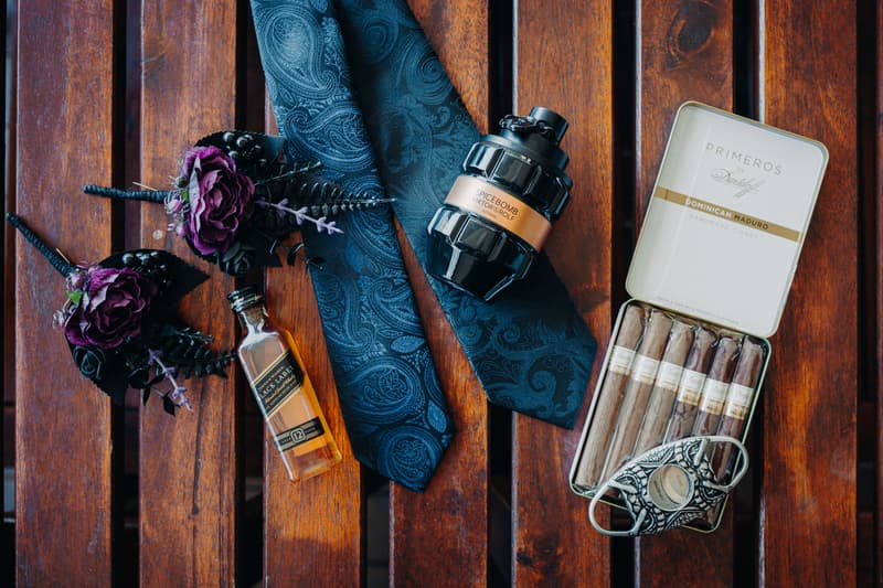 Groom's accessories including two boutonnieres with purple flowers, a paisley-patterned blue tie, a bottle of Johnnie Walker Black Label whiskey, Spicebomb cologne by Viktor & Rolf, and a tin of Primeros Dominican Maduro cigars with a cigar cutter, arranged on a wooden surface at Ocean View Estates.