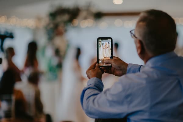 A guest in a blue shirt takes a photo with a smartphone of two brides standing together in wedding dresses in front of a floral arch, with other guests seated and standing nearby.