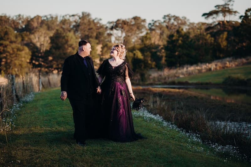 Renae and Adam walk hand in hand along a grassy path beside a lake at Ocean View Estates, with trees in the background during a couple portrait session.