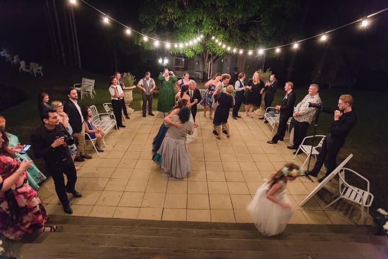 Wedding guests mingle and dance on an outdoor tiled patio at Kwila Lodge during the reception stage, with string lights overhead and a photographer capturing moments.
