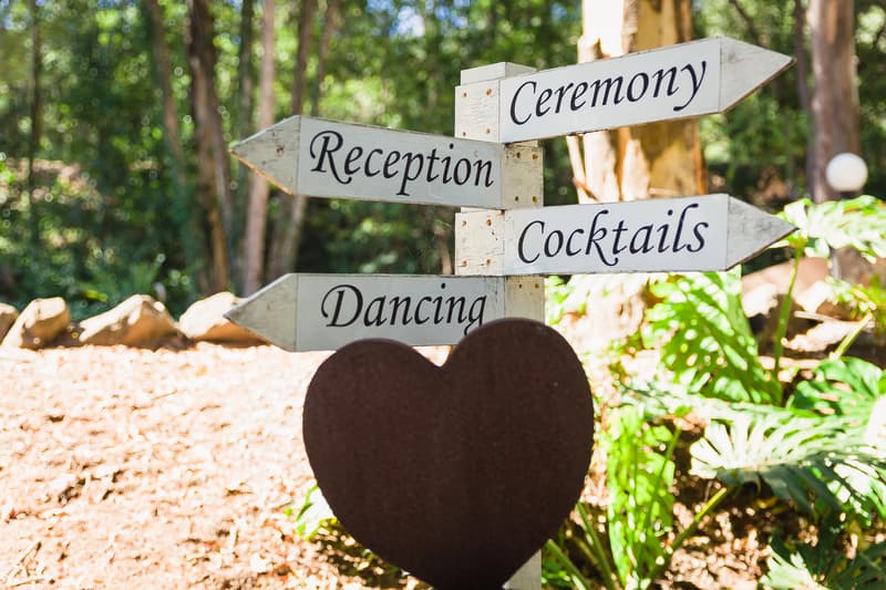 Directional wooden signpost at Kwila Lodge showing arrows for Ceremony, Reception, Cocktails, and Dancing with a heart-shaped decoration in front.