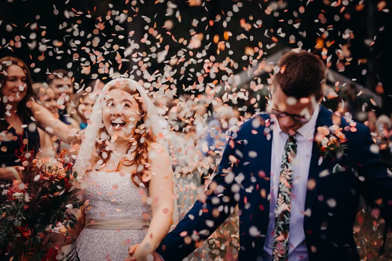 Bride Lilly and groom Connor walk through falling confetti at the ceremony stage at Yabbaloumba Retreat — By The River, surrounded by guests.