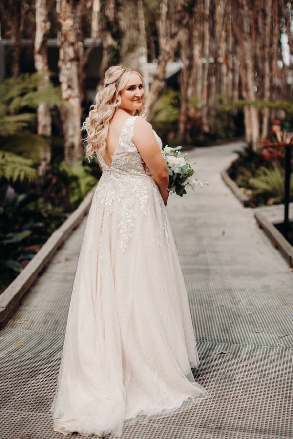 The bride Chloe stands alone on a pathway surrounded by trees and greenery at Sandstone Point Hotel, holding a bouquet and looking back over her shoulder.