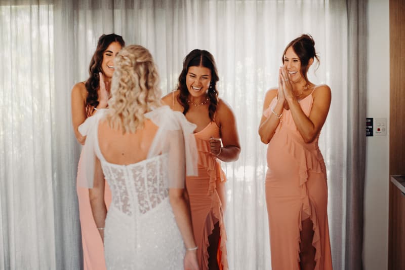 The bride Krystal, seen from behind in her wedding dress, stands facing three bridesmaids in matching peach dresses who are smiling and clapping inside a room with sheer curtains at Sandstone Point Hotel.