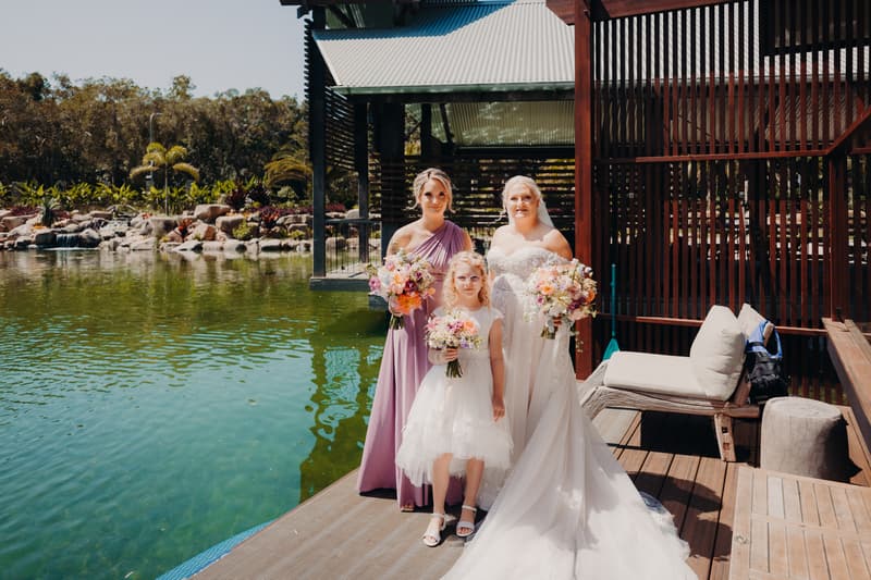 The bride Chantelle, a bridesmaid, and a flower girl pose with bouquets on a wooden deck beside a pond at Sandstone Point Hotel.