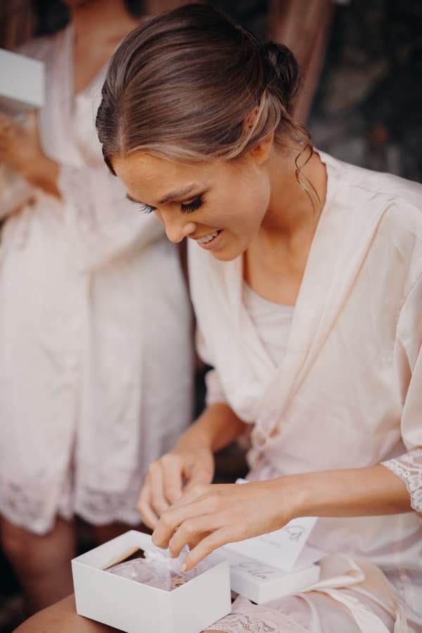 The bride Chelsea, wearing a light pink robe, is seated and opening a white gift box during bridal preparations at Preston Peak Winery Functions Centre.