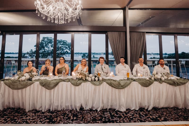 The bride Chloe and groom Brodie sit at the center of a long head table at Sandstone Point Hotel — Pumicestone Room, flanked by bridesmaids and groomsmen, with floral arrangements and candles decorating the table and large windows showing an outdoor waterfront view behind them.