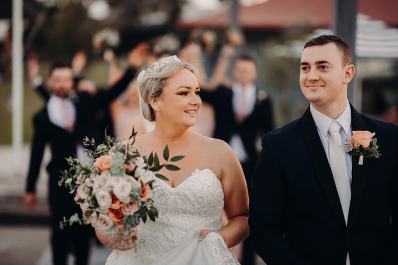 Emily the bride and Dylan the groom walk together outdoors with the bridal party blurred in the background at Sandstone Point Hotel.