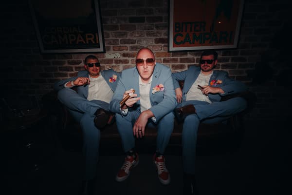 Three men in matching light blue suits with boutonnieres and sunglasses sit on a leather couch, each holding a cigar. Wedding photographers Brisbane by She Said Yes.