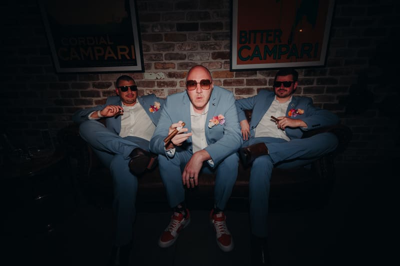 Three men in matching light blue suits with boutonnieres and sunglasses sit on a leather couch, each holding a cigar. Wedding photographers Brisbane by She Said Yes.