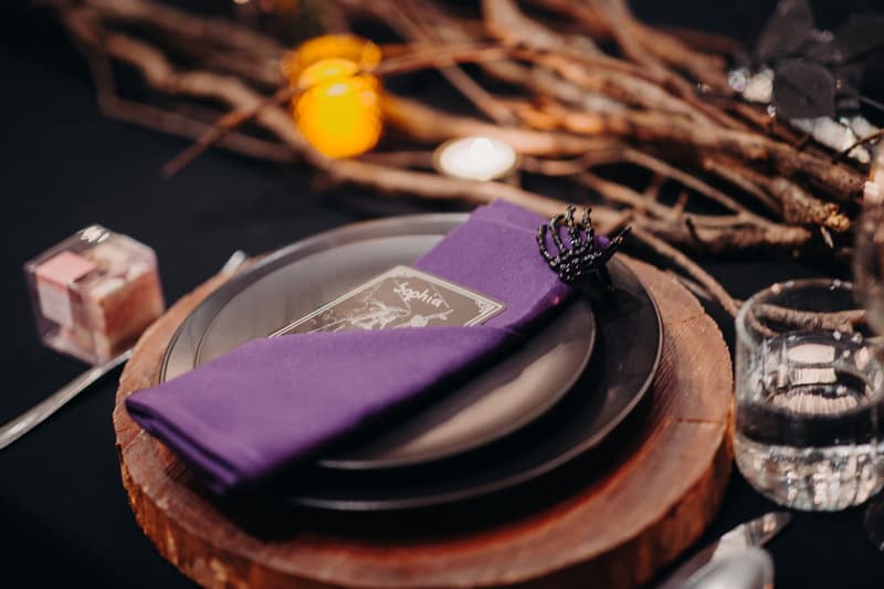 Close-up of a wedding reception table setting at Ocean View Estates — Function Room featuring a wooden charger plate, stacked black plates, a purple napkin with a black decorative ring, a place card, a glass of water, and a small candle in the background.