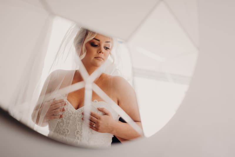 The bride Emily adjusts her wedding dress, seen through a decorative mirror reflection.