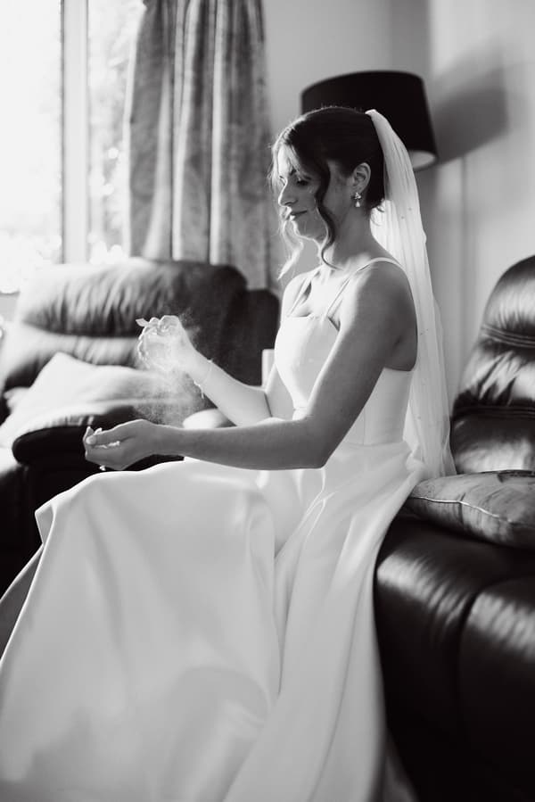 The bride Ashleigh sits on a leather couch at Sandstone Point Hotel, applying perfume to her wrist while wearing her wedding dress and veil.
