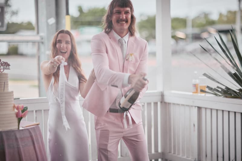 The bride Olivia in a white dress and the groom Jake in a pink suit are on the reception stage at White Horse Ranch. Jake is opening a bottle of champagne while Olivia holds a ribbon and smiles. A wedding cake is partially visible on a table to the left.