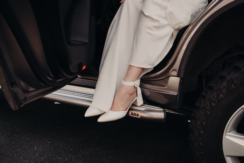 The bride's legs and white shoes are visible as she sits in an open car door, wearing a white wedding dress.