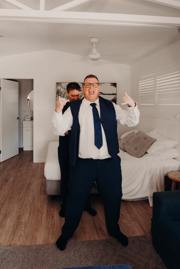 The groom stands in a room at Sandstone Point Hotel, wearing a white shirt, blue tie, and blue vest, making hand gestures while another man adjusts his attire behind him.