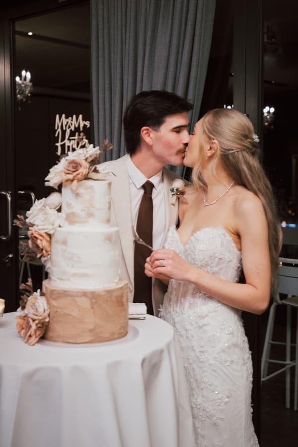 Bride Morgan and groom Joel kiss beside their three-tier wedding cake decorated with flowers at Sandstone Point Hotel — The Pumicestone Room reception.