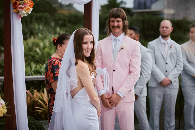 Olivia the bride in a white dress and veil stands smiling at the ceremony stage at Eatons Hill Hotel — Lakeside, with Jake the groom in a light pink suit and groomsmen in light grey suits standing behind him.