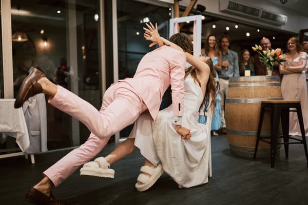 The groom in a pink suit dips the bride in a white dress wearing slippers on the reception stage at White Horse Ranch, while guests watch and smile in the background.