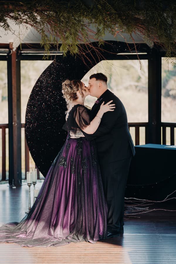 Bride Renae and groom Adam share a kiss on the ceremony stage at Ocean View Estates — On The Lake, standing in front of a large black crescent moon decoration with greenery overhead.