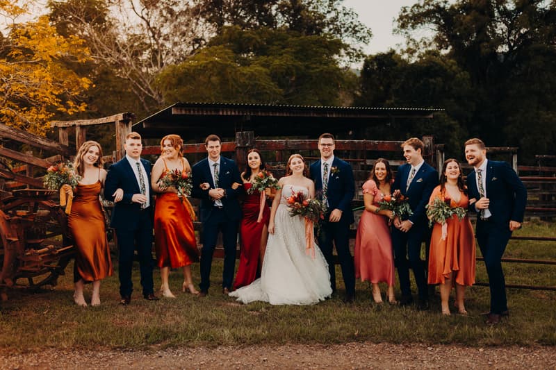 Lilly the bride and Connor the groom stand together with their bridal party at Yabbaloumba Retreat — By The River, all dressed in formal attire with bridesmaids in various shades of orange and red dresses and groomsmen in navy suits, posing outdoors in front of rustic wooden fencing and greenery.
