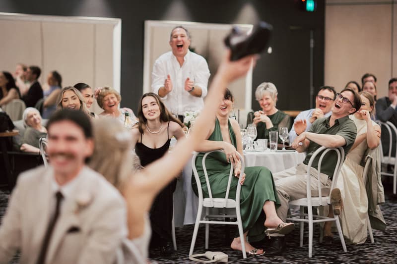 Guests at the Sandstone Point Hotel — The Pumicestone Room reception laugh and applaud while a person in the foreground raises a shoe.