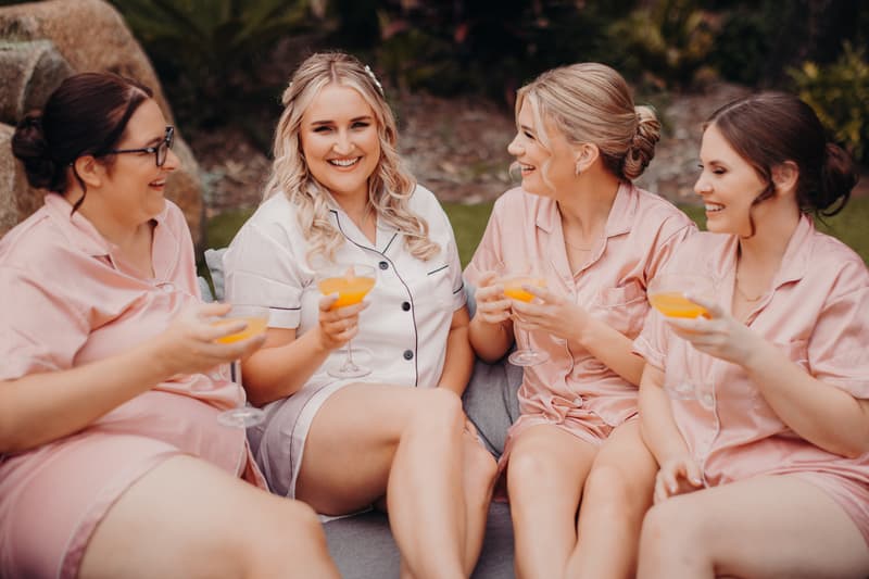 The bride Chloe in white pajamas sits with three bridesmaids in pink pajamas outdoors at Sandstone Point Hotel, holding glasses with orange drinks and smiling.