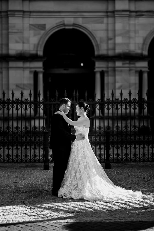 The bride Aria and groom Antony stand facing each other in an embrace outside the Queensland Parliament building, with ornate iron fencing and stone architecture in the background.