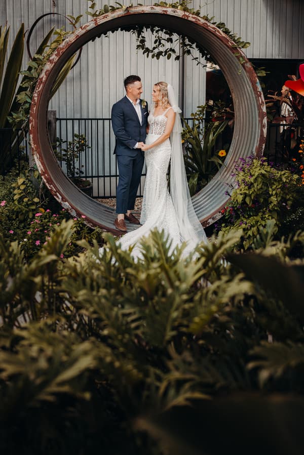 Bride Krystal and groom Brandon stand holding hands inside a large circular metal structure surrounded by plants at Sandstone Point Hotel.