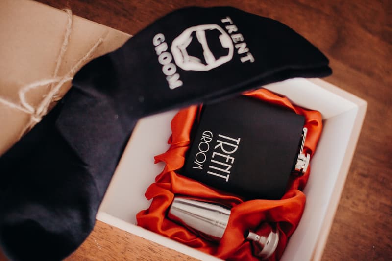 A black flask and a matching black sock with the name 'Trent' and the word 'Groom' printed on both items, placed on a wooden surface with the flask resting on red fabric inside a white box at Ocean View Estates.