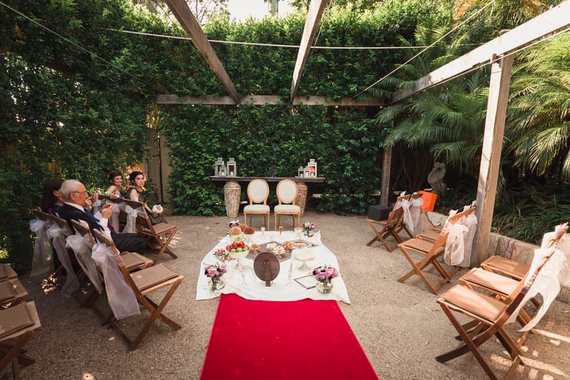 Ceremony setup at Hillstone St Lucia — The Quartyard with two empty chairs at the altar, a red carpet aisle, decorated chairs with white bows, and a low table with floral arrangements and ceremonial items; a few guests seated on the left side.