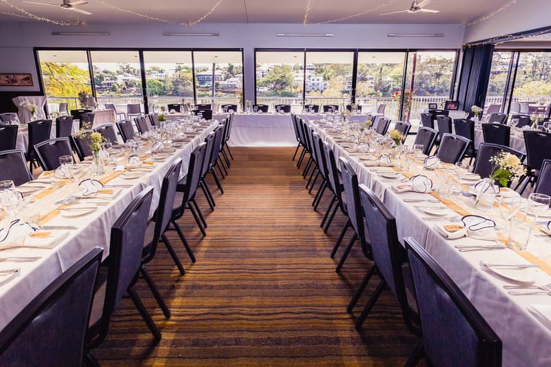 Reception tables set with white tablecloths, glassware, plates, and floral centerpieces in The Malouf Room at Toowong Rowing Club, with large windows showing a riverside view.