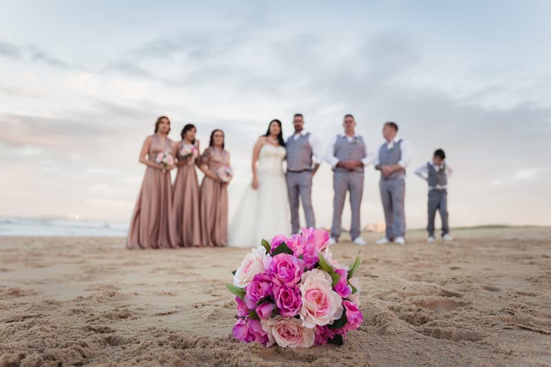 Bride Kelly and groom Shane stand with their bridal party on a sandy beach, with bridesmaids in matching dresses and groomsmen in grey suits behind a bouquet of pink and white flowers placed on the sand.