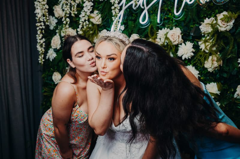 The bride Emily wearing a tiara and white wedding dress poses blowing a kiss while two female guests on either side lean in to kiss her cheeks in front of a floral backdrop at Sandstone Point Hotel — Pumicestone Room.