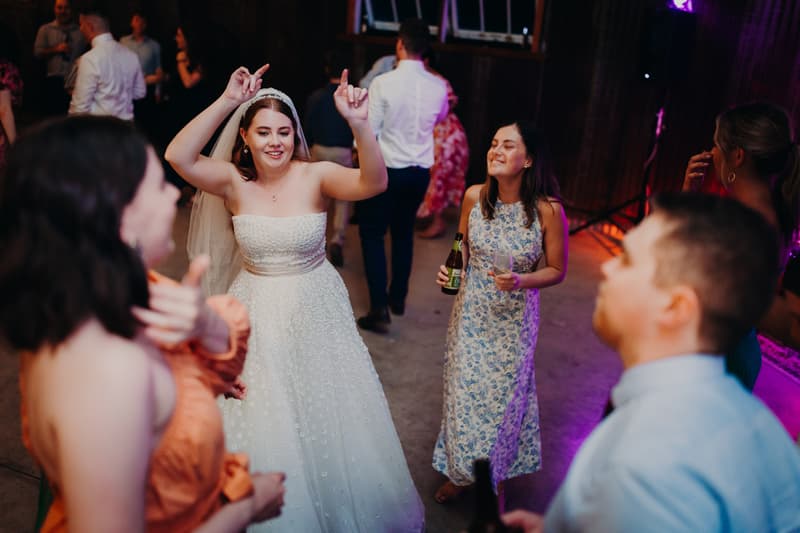 The bride Lilly dances with guests at the reception stage of Yabbaloumba Retreat — The Shed, surrounded by people holding drinks and enjoying the celebration.