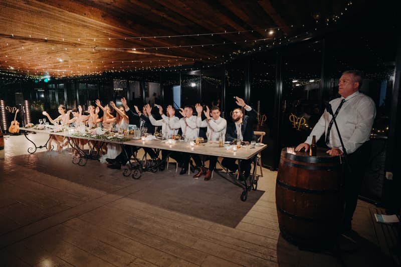 Wedding party seated at a long table at Preston Peak Winery Functions Centre with guests raising their hands, while a man in a white shirt stands at a microphone behind a barrel.